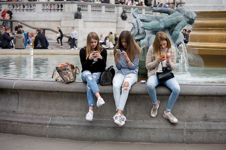Three young women sit on fountain in Trafalgar Square looking at phones