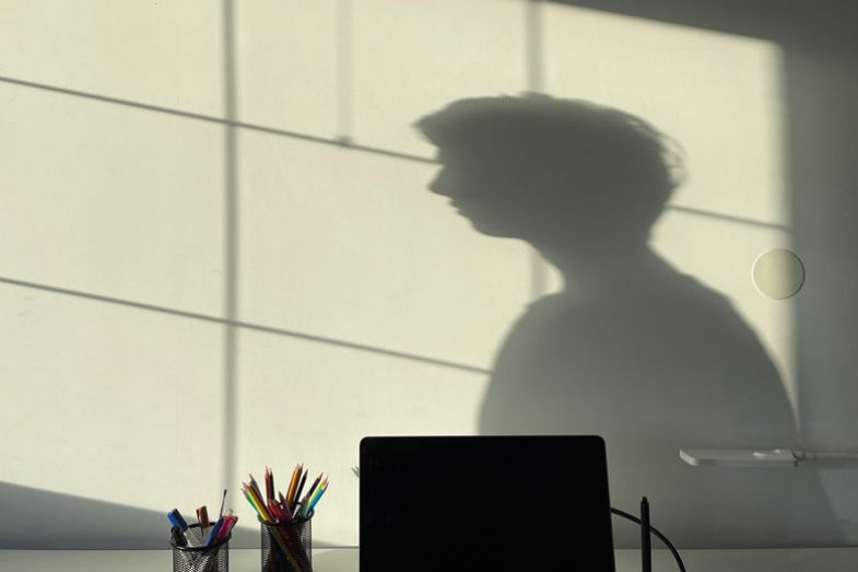 Person shadow on wall desk with laptop and stationary in front as a metaphor for Many students are distressed  and quarantined, unsure how to  finish, how to see people, how to live normally