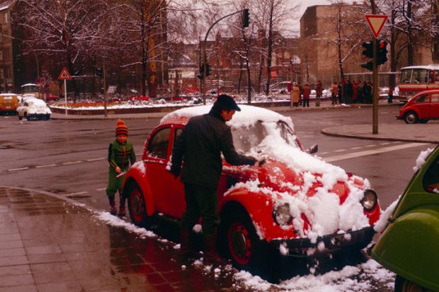 Berlin (West), Germany, 1977. Together, father and son remove snow from the family vehicle at a crossroads. Under the snow, a red VW Beetle hides.