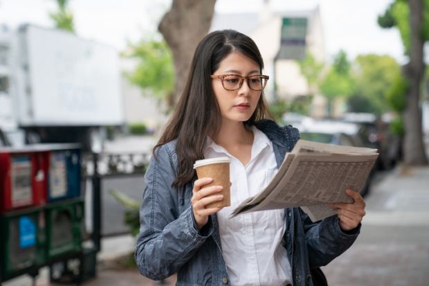 A young woman in the street holds a coffee cup in one hand and a newspaper in the other
