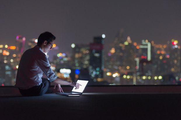 A man in a dark room overlooking a cityscape at night uses a laptop