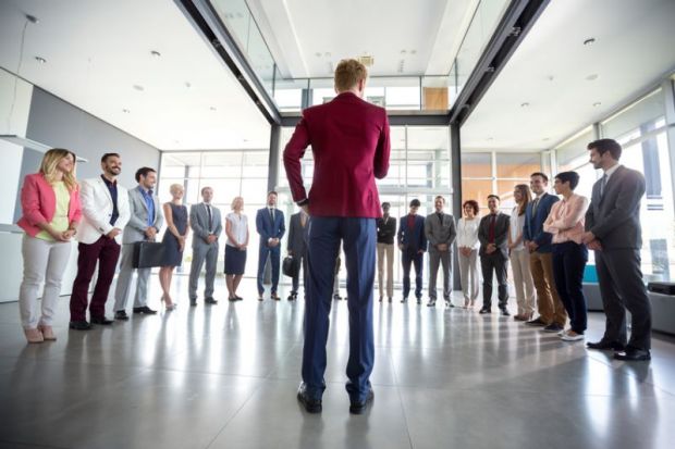 Leadership exemplified by a person in a red blazer addressing a group of diverse professionals gathered in a semicircle, in a bright modern office setting