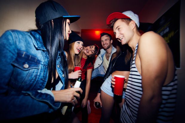 Low angle group of young stylish people standing in dark hall of nightclub together, talking, laughing and drinking beer as they wait to be admitted