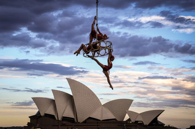Trapeze performers hang from a cable in front of the Sydney Opera House