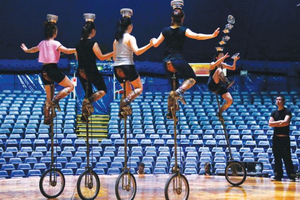 five people on unicycles balancing bowls on their heads with one toppling down as a metaphor for enrolments uncertainty in Australian Universities 
