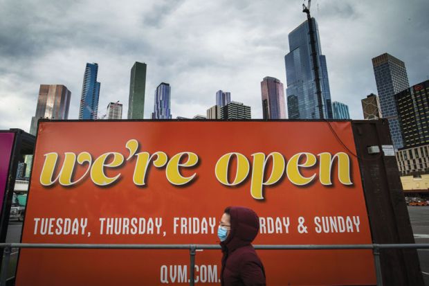 A woman wearing a face mask walks past a billboard reading 'We're open' in Melbourne, Australia to illustrate Australian universities head back to campus after shutdowns