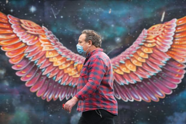 Man with a mask walks in front of wings painting on wall as a metaphor for Which Covid innovations should be embraced 