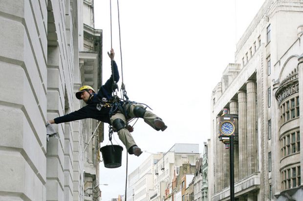 Image of a man suspended from ropes cleans an office in central London as metaphor for that universities in London will be tipped into deficit or pushed further into the red