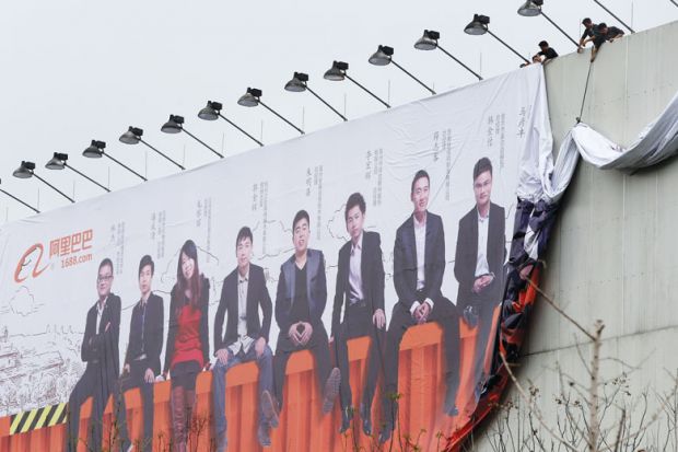 Two people setting up a large billboard of an image of a row of people sitting in a line at the Alibaba Group headquarters for Students seeing little benefit from Chinese start-up drive