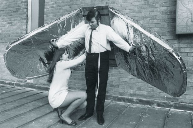 Woman helps a man who prepares for an attempt to fly, using wings made from balsa wood