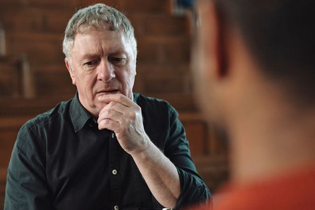Man with grey hair and black shirt holding hand at mouth whilst thinking and talking to male