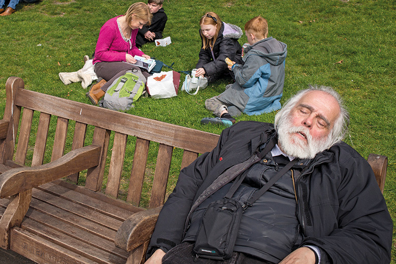 Tramp on park bench with family picnic behind